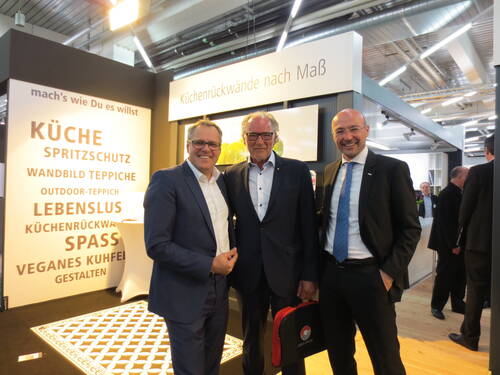 Drei lächelnde Männer im Anzug stehen auf einer Messe vor einem Stand für maßgefertigte Küchenrückwände und Küchenausstattung.