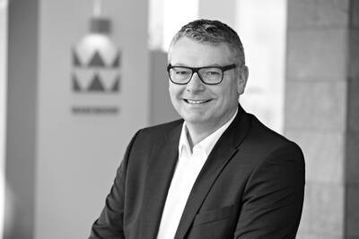 Lächelnder Mann im Anzug mit Brille in einem modernen Büro vor unscharfem Firmenlogo