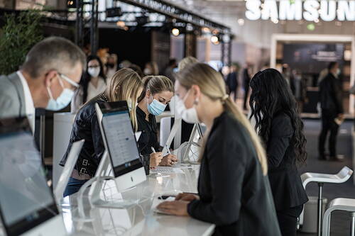 Menschen mit Mund-Nasen-Schutz registrieren sich an Computern an einem Messestand in einer modernen, gut besuchten Ausstellungshalle