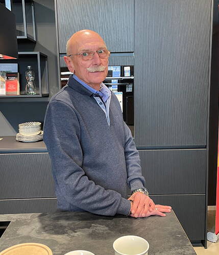 Älterer Mann mit Brille und grauem Pullover lehnt lächelnd an einer modernen, dunklen Küchenzeile