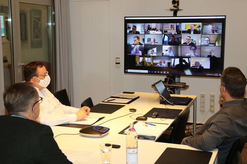 Mehrere maskentragende Personen sitzen in einem Konferenzraum an einem Tisch und nehmen mit Laptops und Unterlagen an einer großen Videokonferenz auf einem Bildschirm teil.
