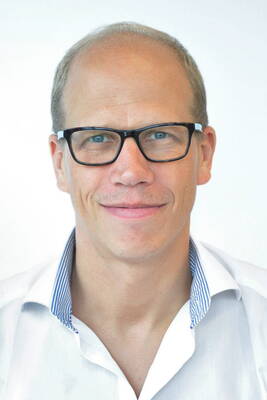 Mittelalterlicher Mann mit Brille lächelt freundlich in die Kamera und trägt ein weißes Hemd mit blau gestreiftem Innenkragen vor hellem Hintergrund.