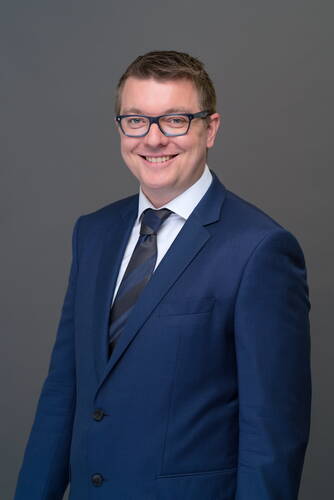 Lächelnder Mann mit Brille im dunkelblauen Business-Anzug vor neutral grauem Hintergrund