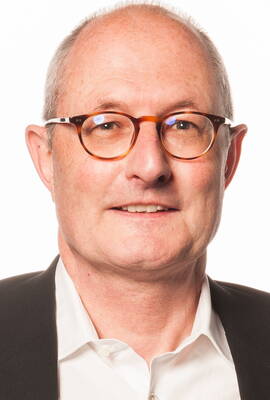 Älterer, glatzköpfiger Mann mit brauner Rundbrille, weißem Hemd und dunklem Sakko blickt freundlich in die Kamera vor hellem Hintergrund.