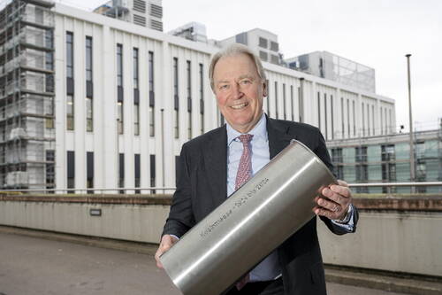 Älterer Mann im Anzug lächelt und hält eine große, silberne Metallröhre mit eingravierter Aufschrift vor einem modernen Bürogebäude im Hintergrund