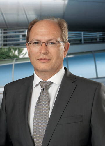 Älterer Mann im grauen Anzug mit Krawatte und Brille blickt ernst in die Kamera vor einem modernen Bürogebäude mit Glasfassade.