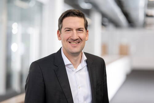 Lächelnder Mann im dunklen Anzug und weißem Hemd steht in einem modernen, hellen Büroflur.