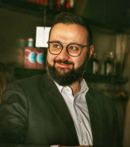 Bartträgtiger Mann mit Brille in elegantem Anzug lächelt und blickt zur Seite in einer stimmungsvoll beleuchteten Bar.