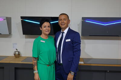 Zwei Messemitarbeitende posieren lächelnd vor modernen Küchenabzugshauben mit blauer LED-Beleuchtung in einem Ausstellungsraum