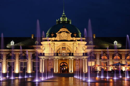 Prunkvolles historisches Gebäude mit beleuchteter Kuppel und barocker Fassade, vor dem bei Nacht farbig angestrahlte Wasserfontänen in die Höhe schießen.