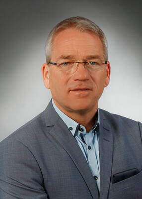 Mittelalterlicher Mann mit grauen Haaren und Brille in hellblauem Hemd und grauem Sakko vor neutralem Studiohintergrund, der ruhig in die Kamera blickt.