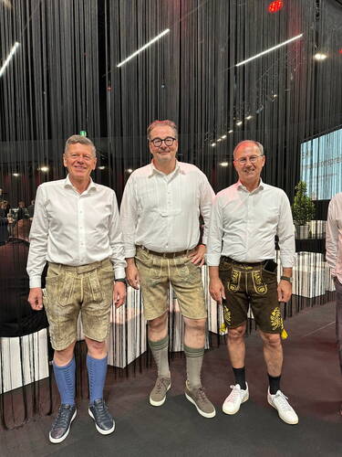 Drei ältere Männer in weißen Hemden und Lederhosen stehen lächelnd nebeneinander in einer modern beleuchteten Halle mit herabhängenden schwarzen Fäden im Hintergrund.