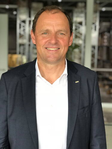 Lächelnder Mann im dunkelblauen Anzug und weißem Hemd steht in einer modernen Industriehalle im Hintergrund