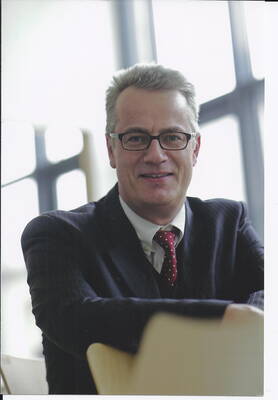 Mittelalterlicher Mann im Anzug mit roter Krawatte und Brille sitzt lächelnd in einem modernen, lichtdurchfluteten Büro.