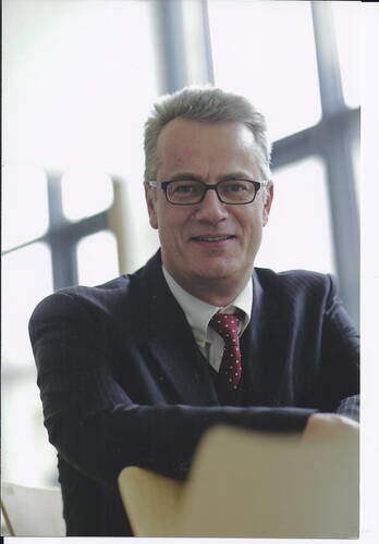 Mittelalterlicher Mann im Anzug mit roter Krawatte und Brille sitzt lächelnd in einem modernen, lichtdurchfluteten Büro.