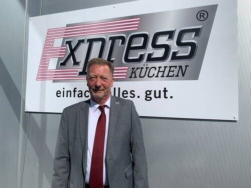 Mittelalterlicher Mann im grauen Anzug mit roter Krawatte steht lächelnd vor dem Firmenlogo „xpress Küchen – einfach. alles. gut.“ an einer Metallfassade