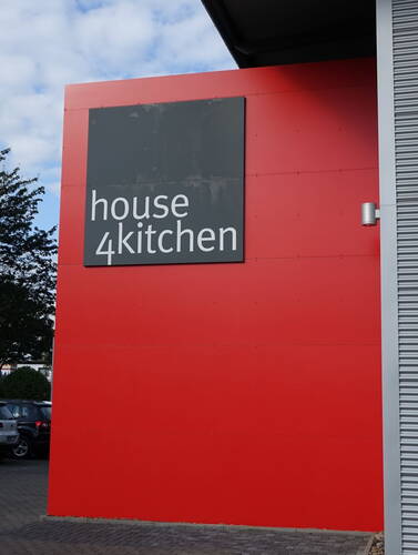 Große rote Gebäudefassade mit grauem Schild „house 4kitchen“ über einem Parkplatz