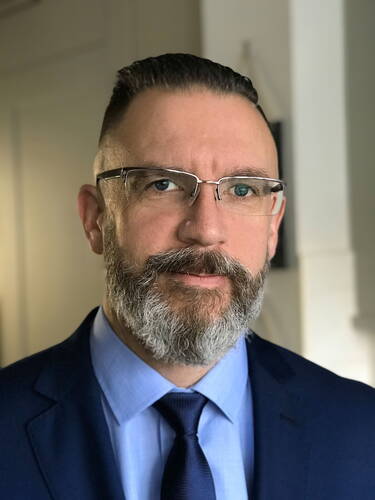 Mittelalterlicher Mann mit grauem Vollbart, Brille und dunkelblauem Anzug blickt ernst in die Kamera in einer hellen Innenraumszene.