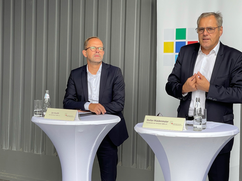 Zwei Geschäftsleute im Anzug stehen an hohen Stehtischen mit Namensschildern und Wasserflaschen und sprechen vor einer grauen Wand mit bunter Logo-Tafel bei einer formellen Veranstaltung