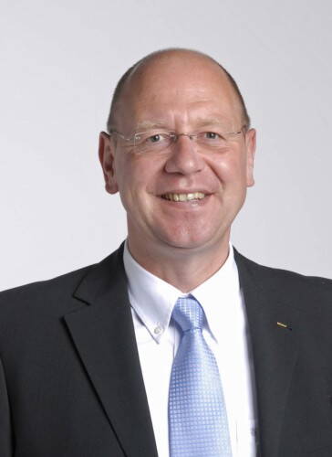 Älterer, lächelnder Mann mit Brille im dunklen Anzug und hellblauer Krawatte vor neutralem Hintergrund