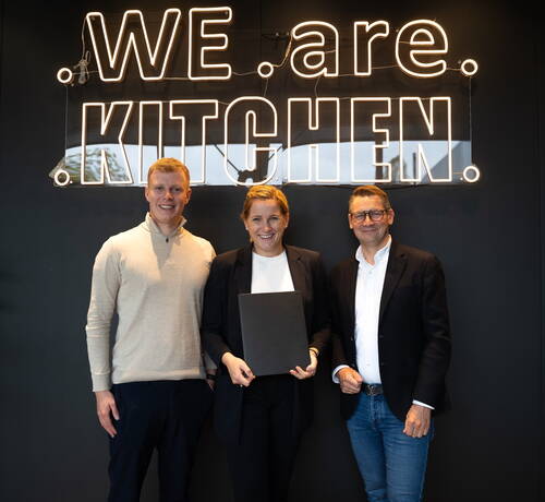 Drei lächelnde Personen stehen vor einer schwarzen Wand mit leuchtendem Schriftzug „WE are KITCHEN“, wobei die Frau in der Mitte eine schwarze Mappe in den Händen hält.