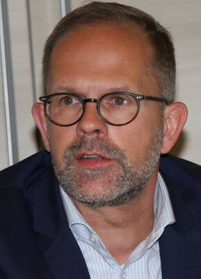 Mittelalterlicher Mann mit Vollbart und runder Brille in Anzug und Hemd spricht konzentriert in einer Innenraumumgebung