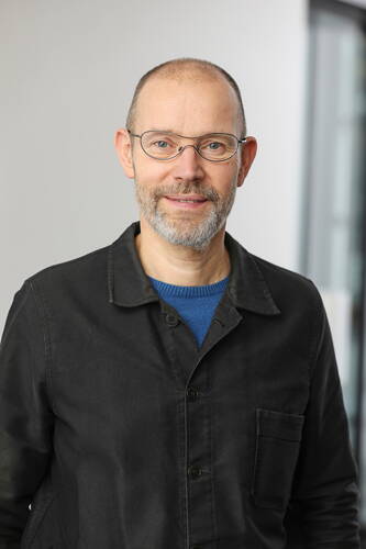 Glatzköpfiger Mann mit Brille und grauem Bart lächelt in die Kamera und trägt eine schwarze Jacke über einem blauen Pullover vor neutralem Hintergrund.
