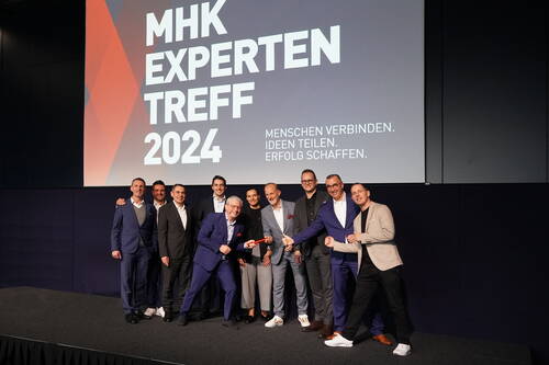 Gruppe von Geschäftsleuten posiert lächelnd auf der Bühne des „MHK Expertentreff 2024“ vor großer Leinwand mit dem Motto „Menschen verbinden. Ideen teilen. Erfolg schaffen.“