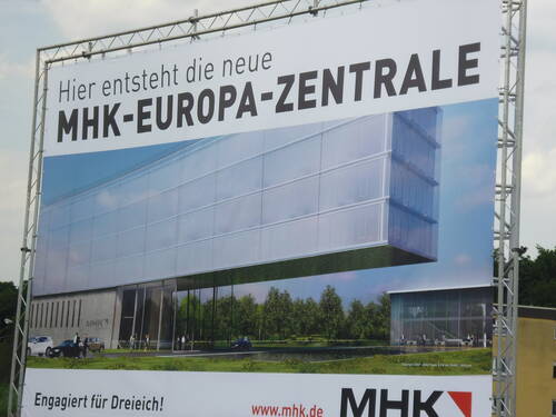 Großes Bauschild mit Visualisierung der geplanten MHK-Europazentrale, das einen modernen gläsernen Bürokomplex mit schwebendem Gebäudeteil, Personen und parkenden Autos in einer grünen Umgebung zeigt.