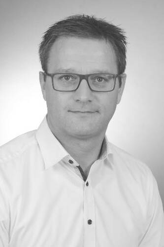 Mittelälterer Mann mit Brille in weißem Hemd blickt ernst und frontal in die Kamera vor neutralem Hintergrund.