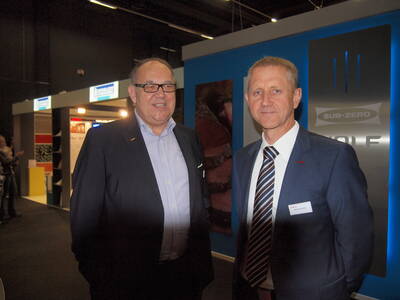 Zwei Geschäftsleute im Anzug lächeln in einer Messehalle vor einem blauen Stand mit dem Logo „Sub-Zero Wolf“ in der Nähe weiterer Ausstellerstände.