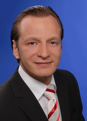 Junger Mann im Anzug mit weiß-rot gestreifter Krawatte lächelt vor blauem Studiohintergrund in die Kamera