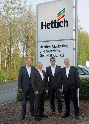 Vier Männer in Anzügen stehen vor dem Firmenschild von Hettich Marketing- und Vertriebs GmbH & Co. KG an einer Einfahrt mit Bäumen im Hintergrund