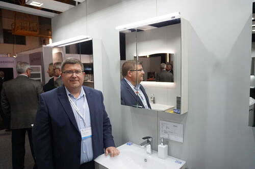 Geschäftsmann im Anzug präsentiert auf einer Messe ein modernes Badmöbel mit beleuchtetem Spiegelschrank und Waschbecken.