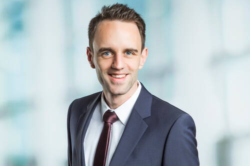 Junger Mann im Businessanzug mit Krawatte lächelt freundlich in die Kamera vor unscharfem, hellem Hintergrund