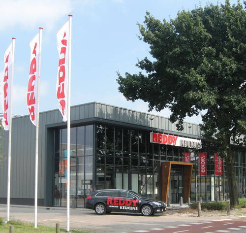 Modernes Küchenstudio von Reddy Keukens mit großer Glasfassade, parkendem Firmenauto und hohen Fahnenmasten an einer Straße neben einem großen Baum