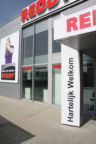 Eingang eines Reddy-Küchenstudios mit großem „Hartelijk Welkom“-Schild und Werbeplakat einer lächelnden Frau, die den Daumen hochzeigt