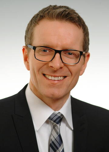 Lächelnder Mann mit Brille im dunklen Anzug und kariertem Schlips vor hellem Hintergrund