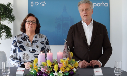 Zwei Personen stehen an einem Tisch mit einem bunten Adventskranz mit vier lila Kerzen und weihnachtlicher Dekoration, im Hintergrund ein blaues porta‑Logo.