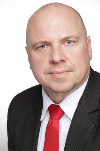 Glatzköpfiger Mann im dunklen Nadelstreifenanzug mit weißem Hemd und roter Krawatte blickt mit neutralem Gesichtsausdruck direkt in die Kamera vor weißem Hintergrund.