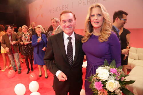 Elegant gekleideter Mann im Anzug und blonde Frau im lilafarbenen Kleid mit Blumenstrauß posieren lächelnd auf rotem Teppich vor feiernder Menschenmenge in festlich dekoriertem Saal.