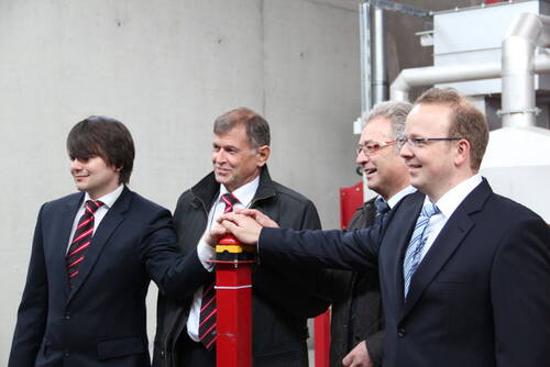 Vier Männer im Anzug drücken gemeinsam einen großen roten Startknopf in einer Industrieanlage und lächeln dabei feierlich.