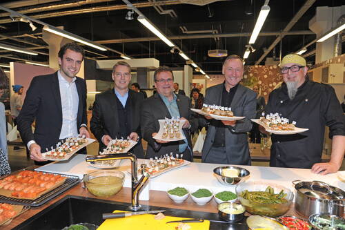 Gruppe von Männern in eleganter Kleidung und Koch im Küchenstudio, die lachend große Platten mit aufwendig angerichteten Fingerfoods präsentieren, während vor ihnen frische Zutaten und Kochutensilien auf der Arbeitsfläche liegen.