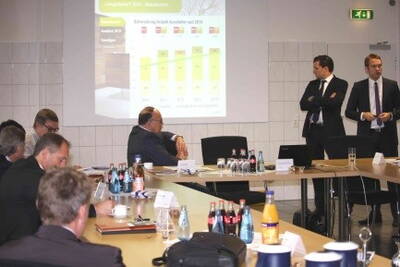 Teilnehmer einer Geschäftsbesprechung verfolgen eine Präsentation mit Wachstumsdiagramm, während zwei Anzugträger vorn die Zahlen erläutern und Getränke auf dem Konferenztisch stehen.