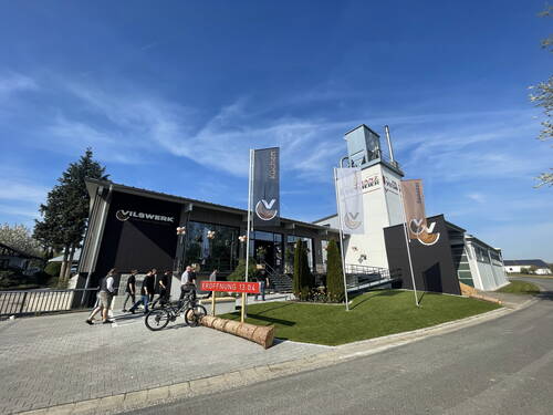 Mehrere Personen betreten bei sonnigem Wetter das modern gestaltete Vilswerk-Gebäude mit Eröffnungsdatum 13.04., umgeben von Fahnen, Grünfläche und einem hohen Turm im Hintergrund.