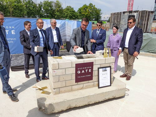 Geschäftsleute in Anzügen setzen gemeinsam einen symbolischen Grundstein für den Bürokomplex „Eschborn Gate“ auf einer Baustelle und halten dabei Ziegelsteine und Maurerkellen bereit