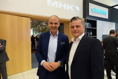 Zwei Geschäftsleute in Anzügen stehen lächelnd in einer Messehalle zwischen den Ständen von MHK Group und Siemens