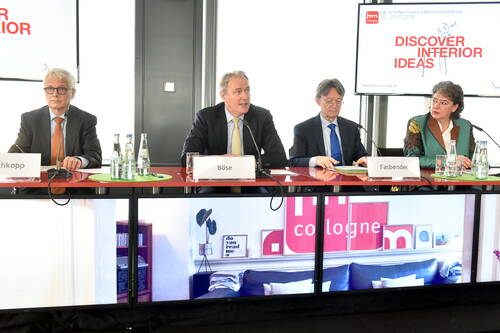 Vier Konferenzteilnehmende sitzen mit Namensschildern und Mikrofonen an einem langen Tisch vor Bildschirmen, während im Hintergrund eine Präsentationsfolie mit der Aufschrift „Discover Interior Ideas“ zur internationalen Einrichtungsmesse in Köln angezeigt wird.