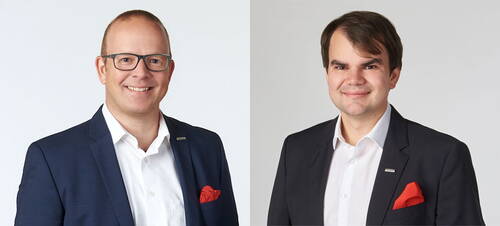 Zwei lächelnde Männer in dunklen Anzügen mit weißen Hemden und roten Einstecktüchern stehen vor hellem Hintergrund nebeneinander in einem professionellen Porträt.