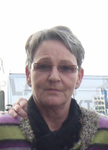 Ältere Frau mit kurzem grauem Haar und Brille blickt ruhig in die Kamera, während eine Hand beruhigend auf ihrer Schulter ruht.